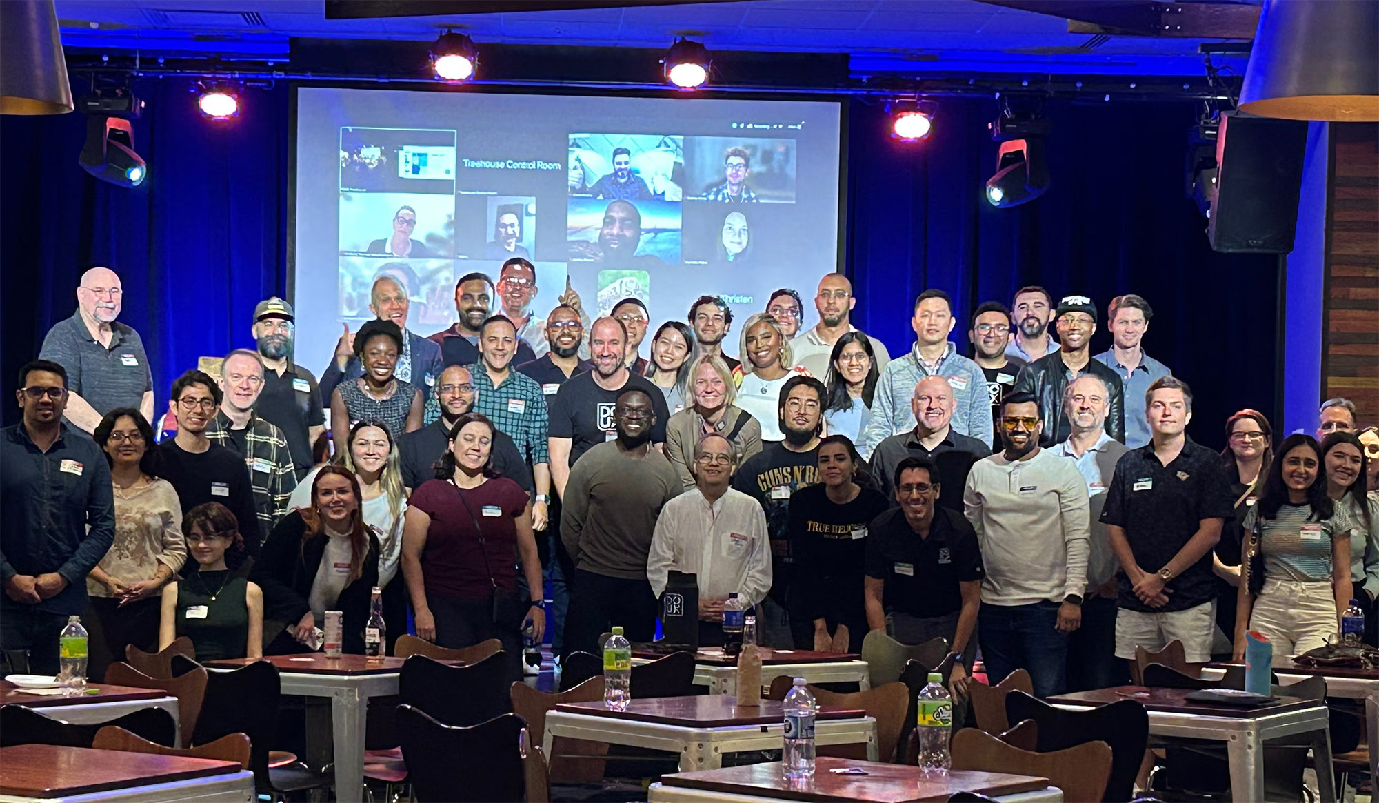 Photo of a group of about 40 people in front of a stage with others on a Zoom screen in the background.